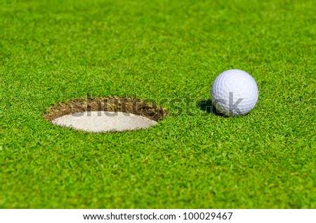 A hole over golf ball on green. Shallow depth of field. Focus on the ball and the hole.