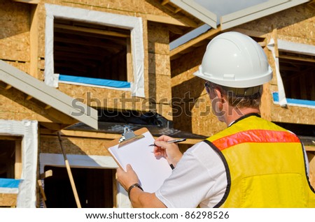 A man in a hard hat standing in front of an house holding a clipboard in his hand.