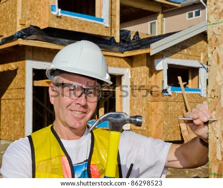 A man in a hard hat standing in front of an house holding a hammer and nail.