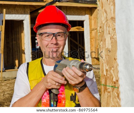 A man in a hard hat standing in front of an house holding a drill in his hand.