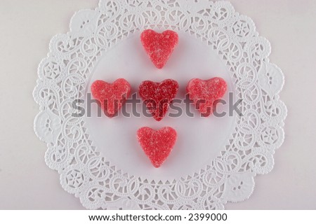 Single red and four pink sugar coated heart candies on a white doily.