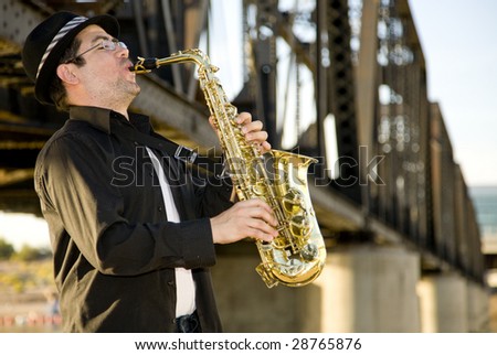 A saxophonist plays outdoors against an  industrial backdrop.
