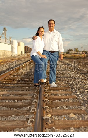 A multicultural couple photographed at sunset in an industrial location  featuring railroad tracks.