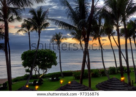 The sun sets on the beach of a resort with grass and lounge chairs in Lahaina, Maui, Hawaii.