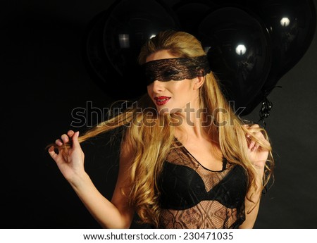 Young beautiful slim girl wearing black lace sensual top and black skirt with evening make up and black lace band on her eyes posing with black balloons and playing with her hair on black background