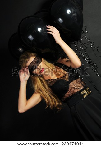 Young beautiful slim girl wearing black lace sensual top and black skirt with evening make up and black lace band on her eyes posing and playing with black balloons on black background