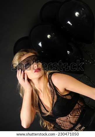 Young beautiful slim girl wearing black lace sensual top and black skirt with evening make up and black lace band on her eyes posing with black balloons and touching her ear on black background