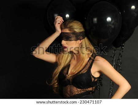 Young beautiful slim girl wearing black lace sensual top and black skirt with evening make up and black lace band on her eyes posing with black balloons looking aside on black background