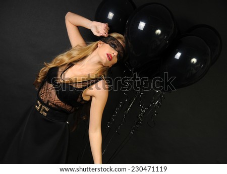 Young beautiful slim girl wearing black lace sensual top and black skirt with evening make up and black lace band on her eyes posing and making sensual pose with black balloons on black background