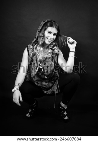 black and White portrait of a beautiful smiling caucasian girl with long curly hair wearing Golden vest with black polka dots playing with her curls seating on the black background