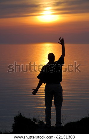 silhouetted Man wave in sunset stand on cliff wave in yellow sunset, smooth water reflecting the sun in horizon, spectacular layered sky. Raised and lowered arm in short sleeve shirt, summer night.