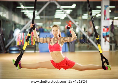 Young attractive woman does splits crossfit stretching with trx fitness straps in the gym\'s studio, shows biceps and smiles