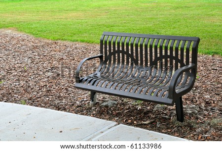 Park bench made of metal. Painted brown and surrounded by concrete, leaves and grass.