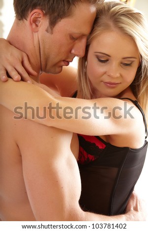 Young and fit caucasian adult couple in an embrace. Semi-nude and topless on a bed in a light bedroom with the woman wearing a sexy red and black lace corset and the man wearing only blue jeans.