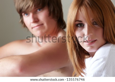 Young adult caucasian man and woman sitting half undressed on a bed. The man is shirtless or topless with blue jeans and the girl is wearing a white cotton robe
