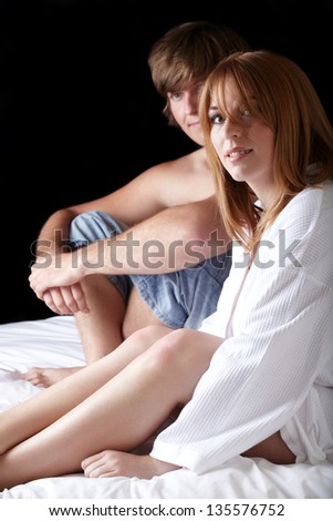 Young adult caucasian man and woman sitting half undressed on a bed. The man is shirtless or topless with blue jeans and the girl is wearing a white cotton robe