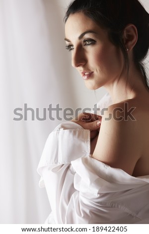Sexy young woman with black hair in a white shirt undressing in front of a window