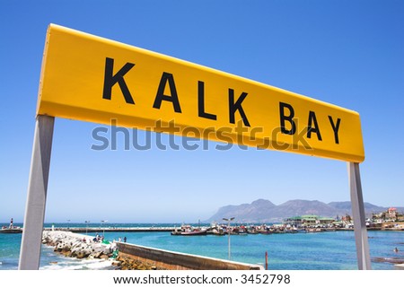Yellow train station sign against a blue sky with the Kalk Bay harbour in the background