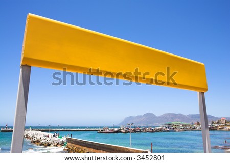 Yellow train station sign against a blue sky with the Kalk Bay harbour in the background