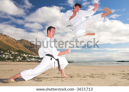 Young adult men with black belt practicing on the beach on a sunny day. The man doing the flying kick in the background has movement. Focus on the standing man