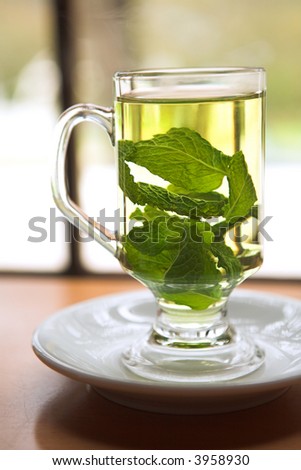Hot green mint tea standing next to a window with whole mint leaves