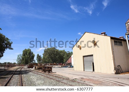 Rusted metal mining wagons standing outside an industrial warehouse with train tracks running in front of the building. Blue sky and sunny day