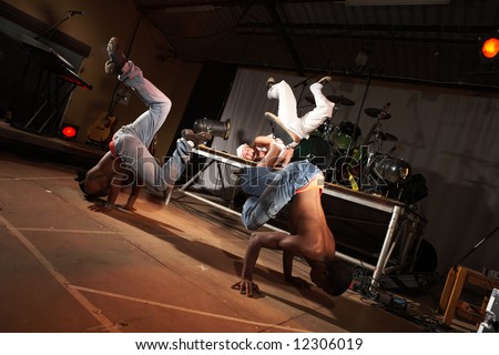 Three freestyle hip-hop dancers in a dancing training session. 2 young adult males and a female in a home training studio with stage and instruments. Lit with spotlights. Movement on edges of dancers