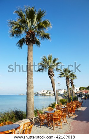 Empty wicker tables and chairs at an outdoor cafe with Tall palm tree against a clear blue sky and the Aegean in the background on a sunny day