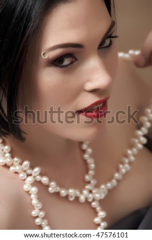 Sexy naked young caucasian adult woman with red lips, short black hair and a pierced eyebrow, covered in a dark satin sheet and lying on a bed, wearing a string of white pearls