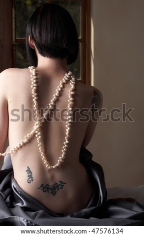 Sexy naked young caucasian adult woman with short black hair and covered in a dark satin sheet, sitting on a bed with her back to the camera and wearing a string of pearls