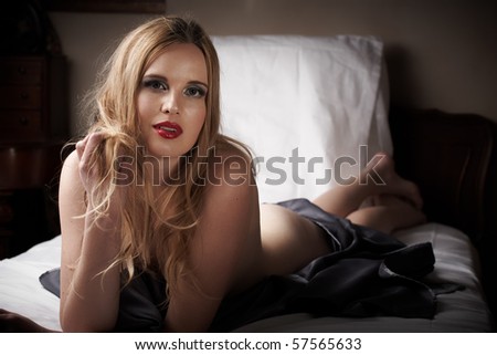 Sexy naked young caucasian adult woman with red lips and blonde hair lying on a bed, covered with a dark silk sheet. Single light source