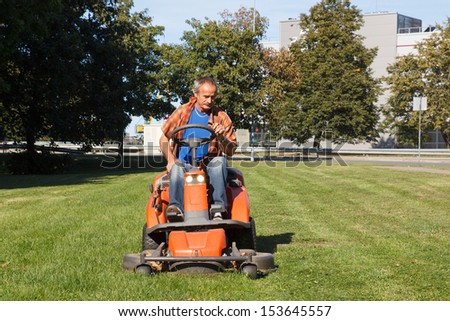 man driving a red lawn mower (tractor) in the city park