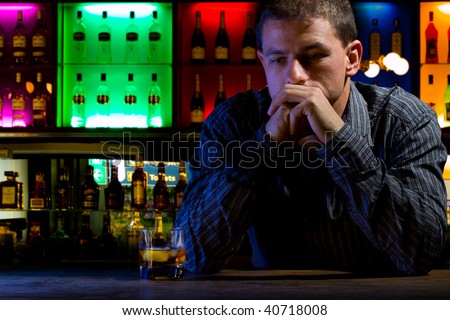 Worried man sitting at bar with whiskey glass. Dark night scene. Colored background