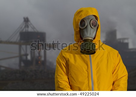 Man wearing gas mask standing infront of factory. Frontal shot. Background out of focus
