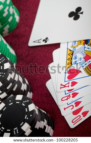 A closeup of a poker hand that just misses getting a Royal Flush. There is straight of hearts-ten, Jack, Queen, King but the Ace is a club with checkered poker chips lined up on the left.