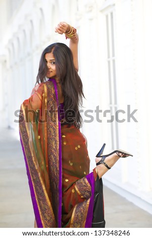 A young, 30 something, Indian woman with long black hair, dressed in traditional purple and gold Indian sari, kicks up her heels while walking down white hallway