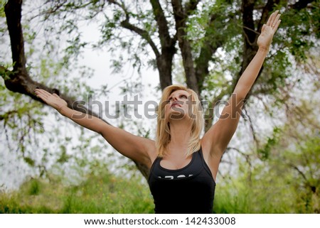 A mature woman wearing black workout clothes stands with her arms outstretched, aligned with the tree branches behind her