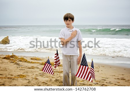 A young boy, with sand all over his hands and pants, plants four American flags into the sand at the beach then stands among them with his hand to his heart