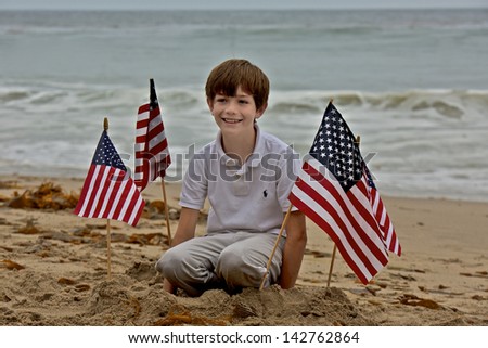 A young boy smiles as he sits among four American flags that be planted in the sand at a beach