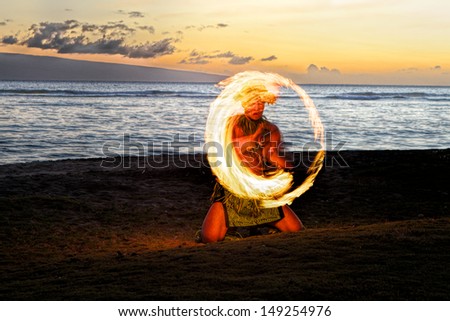 A male fire dancer kneels on a Hawaiian beach at dusk, spins a stick with fire to create a glowing circle of fire in the air.