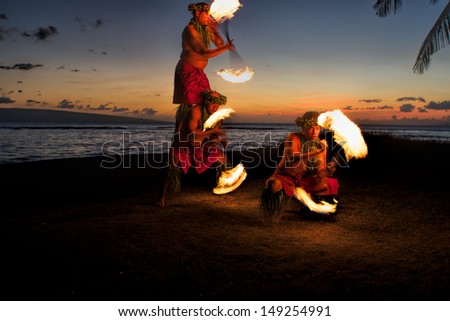 Bathed in an orange glow, three male fire dancers on a Hawaiian beach at dusk, spin sticks with fire on either side, creating patterns in the air. The glow of the fire illuminates the ground.