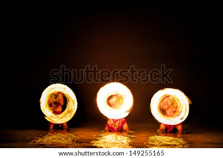 Bathed in an orange glow, three male fire dancers, stand in the ocean while quickly spinning sticks with fire, creating giant circles of fire in the air that are also reflected on the water.