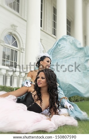 Glamorous young African American woman wearing black bustier and full length puffy pink skirt lies down on grass in front of Asian woman wearing old fashioned dress with its trail blowing in the wind