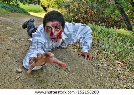 A male zombie crawls along the ground on a dirt path reaching one hand forward