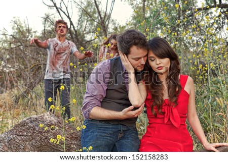 Two grotesque and bloody zombies slowly walk up behind an unsuspecting couple as they cuddle in a city park
