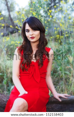 Portrait of a young 20-something woman dressed in a bright red dress, posing in a wooded area, framed by yellow flowers