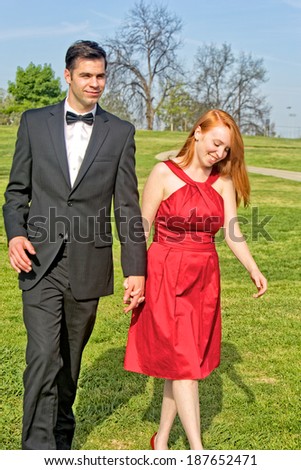 Young man and woman dressed for a special occasion (possibly a wedding), hold hands as they walk through a meadow in a city park.