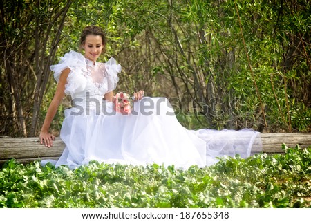 A smiling young bride dressed in a flowery and puffy white wedding dress sits on a wooden log in a grove of trees.