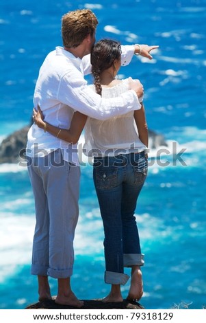 A young couple stands at the top of a cliff overlooking the ocean. The man is pointing to something in the distance. Vertical image orientation.