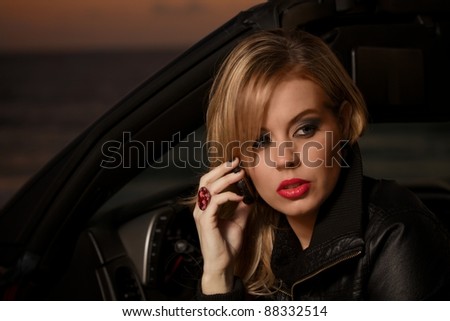 A beautiful blond woman sits in her car, with the door open, and talks on a cell phone in front of the ocean at sunset